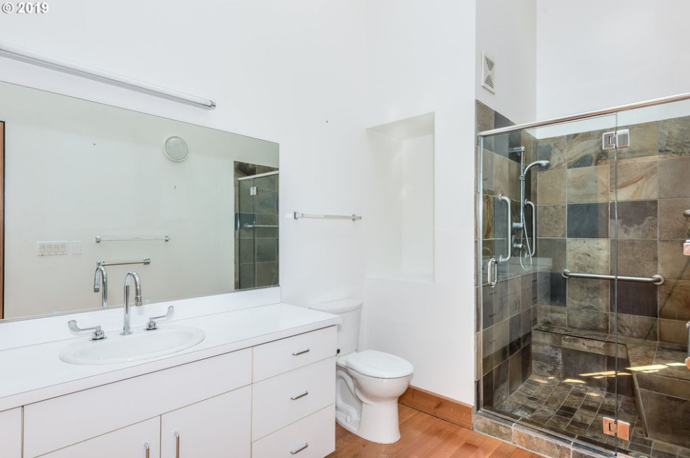 Bathroom - mid-sized industrial master medium tone wood floor, brown floor, single-sink and exposed beam bathroom idea in Portland with flat-panel cabinets, white cabinets, a one-piece toilet, white walls, a drop-in sink, laminate countertops, white countertops and a niche