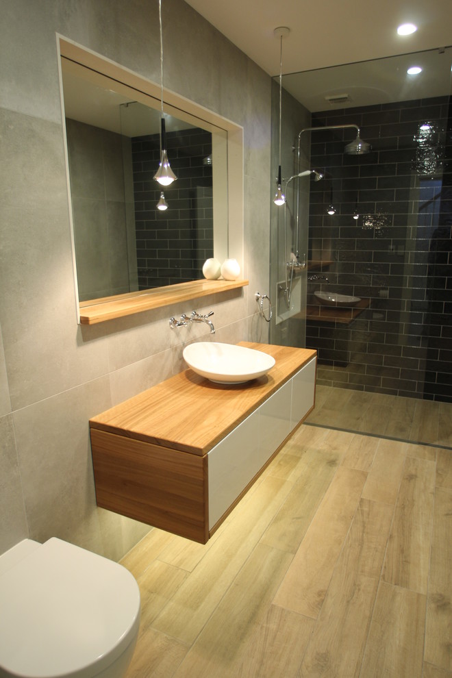 Idéer för mellanstora funkis badrum, med möbel-liknande, skåp i ljust trä, en öppen dusch, en toalettstol med hel cisternkåpa, grå kakel, porslinskakel, grå väggar, klinkergolv i porslin, träbänkskiva, grått golv, med dusch som är öppen och ett fristående handfat
