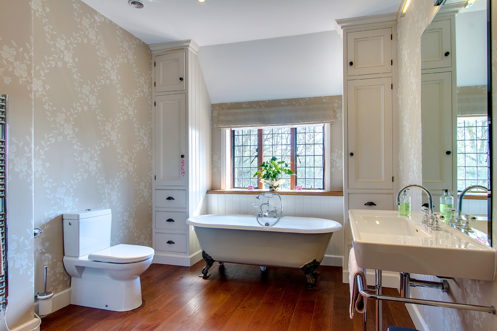 Foto på ett vintage badrum, med ett konsol handfat, vita skåp, ett badkar med tassar, en toalettstol med separat cisternkåpa, beige väggar, mörkt trägolv och luckor med infälld panel