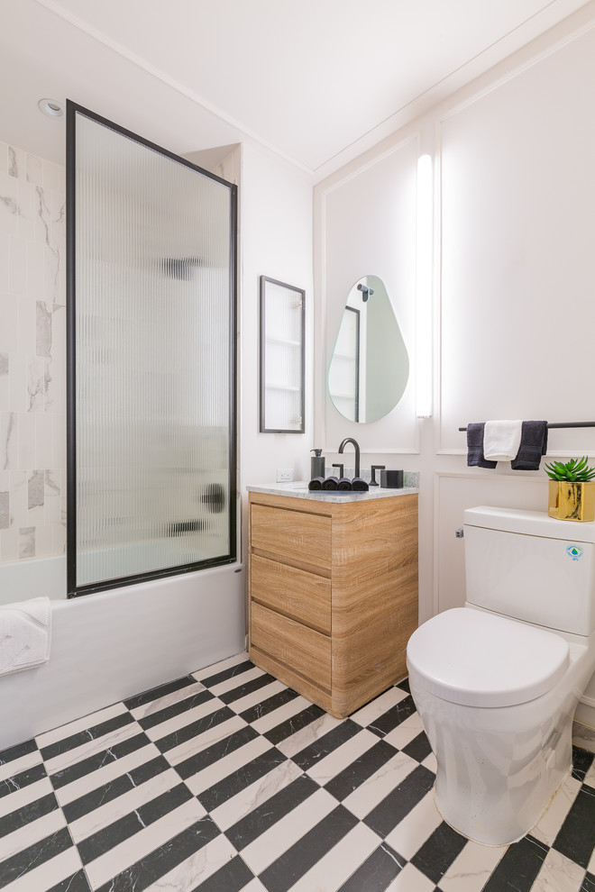Cette image montre une salle d'eau design en bois clair avec un placard à porte plane, une baignoire en alcôve, un combiné douche/baignoire, WC séparés, un mur blanc, un sol en marbre, un lavabo encastré, un sol noir et du carrelage bicolore.