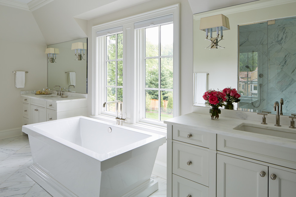 На фото: главная ванная комната в классическом стиле с врезной раковиной, фасадами с утопленной филенкой, белыми фасадами, белыми стенами, отдельно стоящей ванной, каменной плиткой, мраморным полом и мраморной столешницей