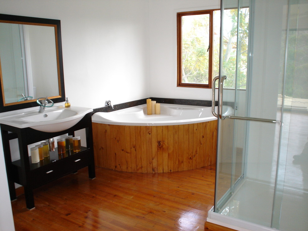 Large coastal ensuite bathroom in Other with a pedestal sink, open cabinets, light wood cabinets, a corner bath, a corner shower, a one-piece toilet, white walls and light hardwood flooring.