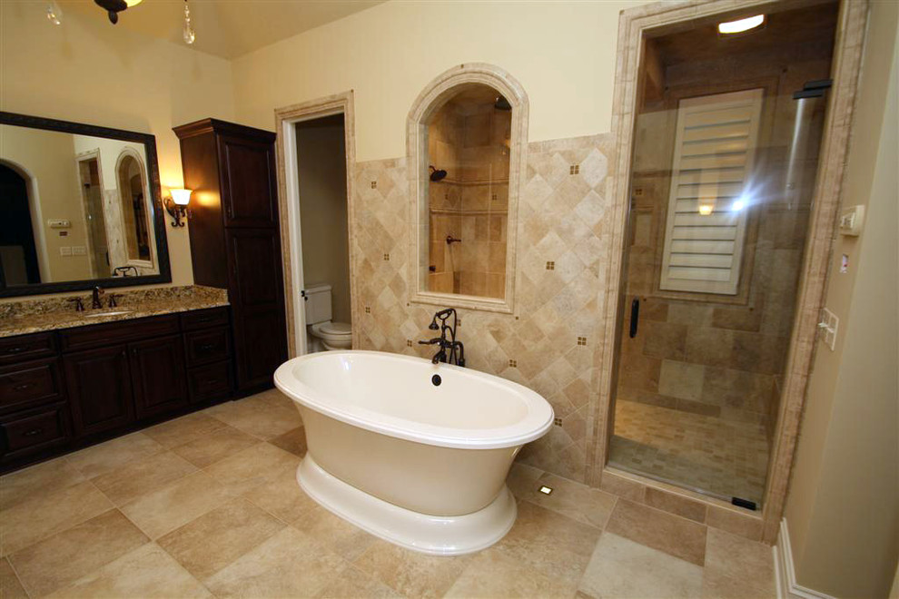 Свежая идея для дизайна: главная ванная комната среднего размера в классическом стиле с фасадами с выступающей филенкой, коричневыми фасадами, отдельно стоящей ванной, двойным душем, бежевой плиткой, каменной плиткой, желтыми стенами, полом из керамогранита, врезной раковиной, столешницей из гранита, бежевым полом и душем с распашными дверями - отличное фото интерьера