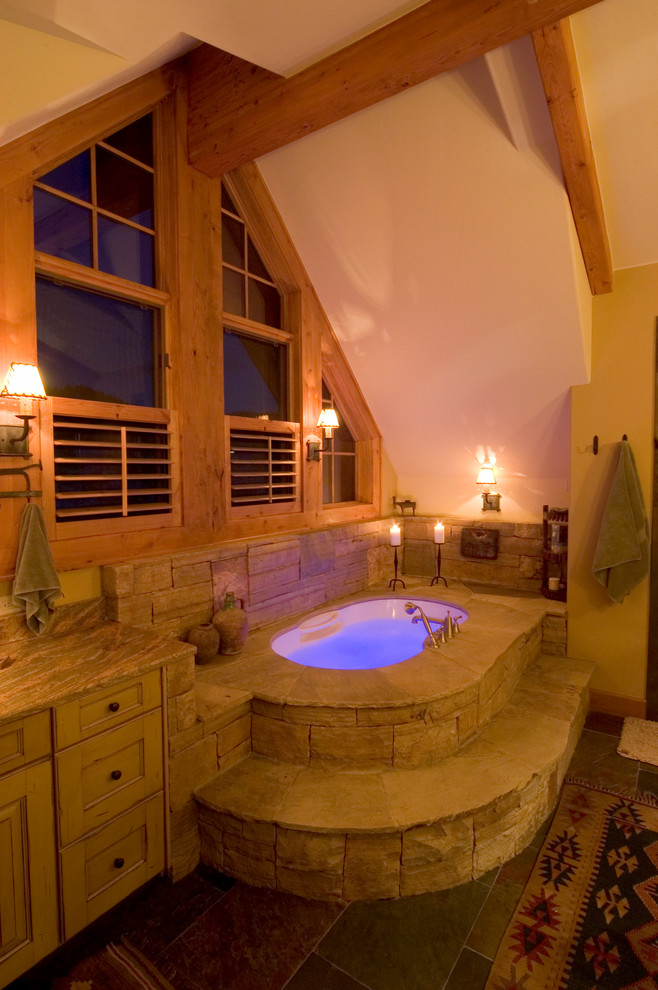 Large rustic ensuite bathroom in Denver with a submerged sink, recessed-panel cabinets, distressed cabinets, granite worktops, a built-in bath, a two-piece toilet, stone tiles, yellow walls and limestone flooring.