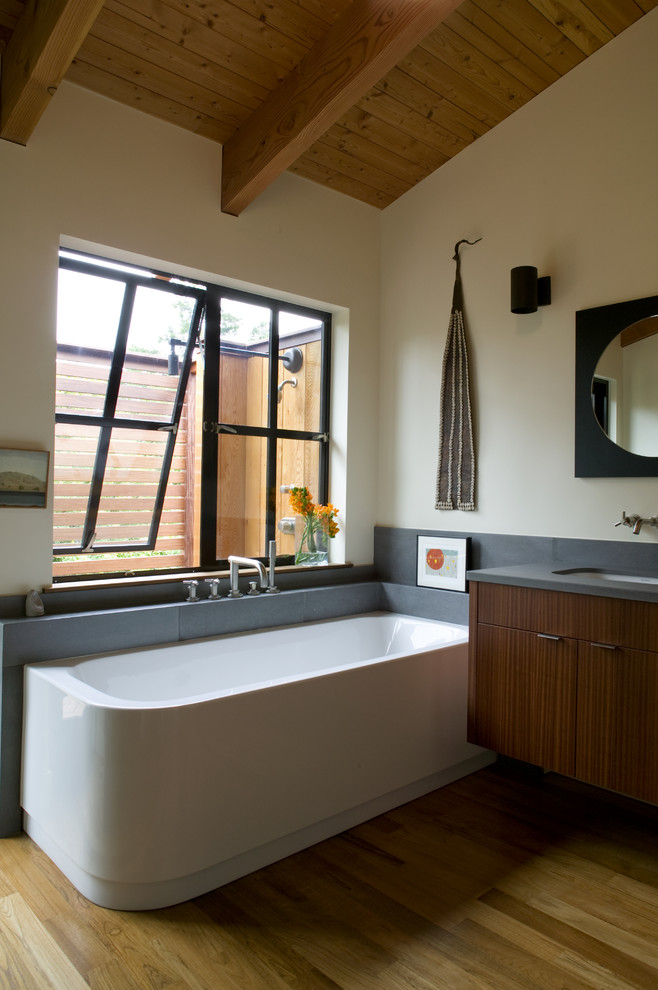 Cette photo montre une grande salle de bain principale chic en bois foncé avec un placard à porte plane, une baignoire d'angle, un carrelage gris, un carrelage de pierre, un mur beige, un sol en bois brun et un lavabo encastré.
