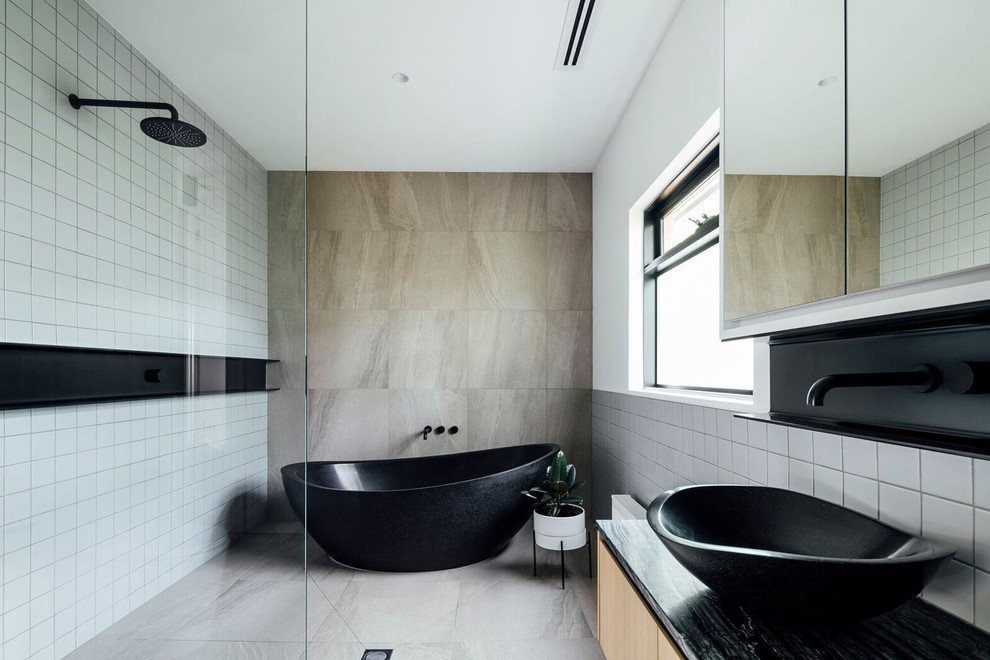 メルボルンにあるコンテンポラリースタイルのおしゃれな浴室 (フラットパネル扉のキャビネット、淡色木目調キャビネット、置き型浴槽、白いタイル、ベッセル式洗面器、グレーの床、黒い洗面カウンター) の写真