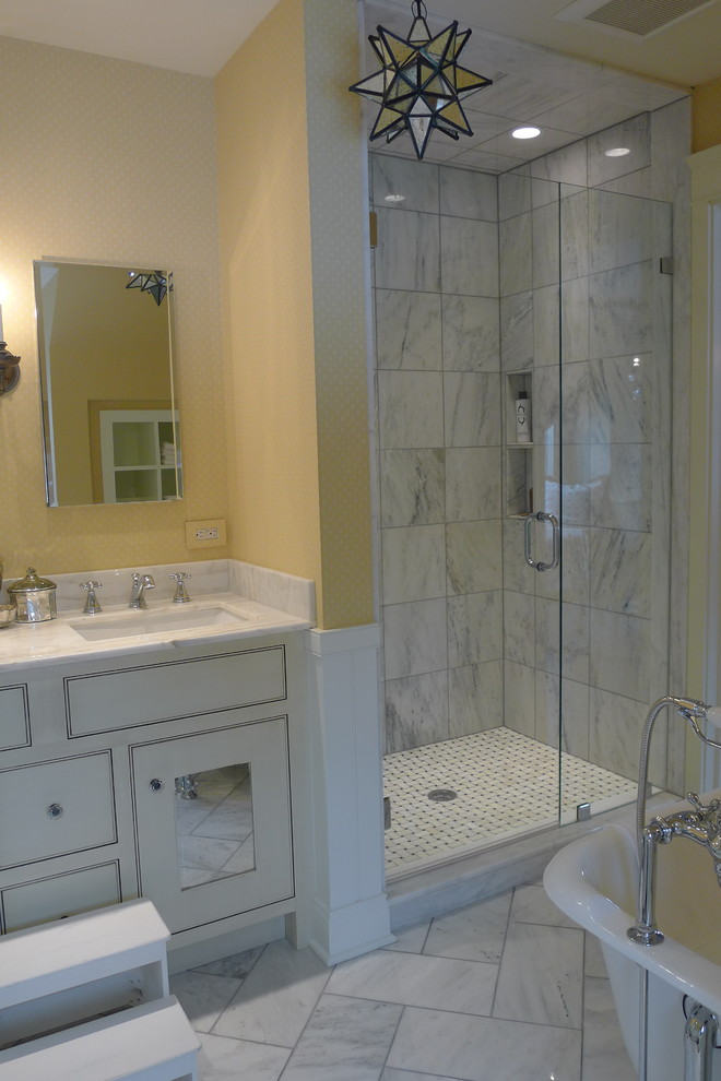 Klassisk inredning av ett litet badrum för barn, med ett undermonterad handfat, vita skåp, marmorbänkskiva, ett badkar med tassar, en dusch i en alkov, en toalettstol med hel cisternkåpa, vit kakel, stenkakel, luckor med profilerade fronter, gula väggar och marmorgolv