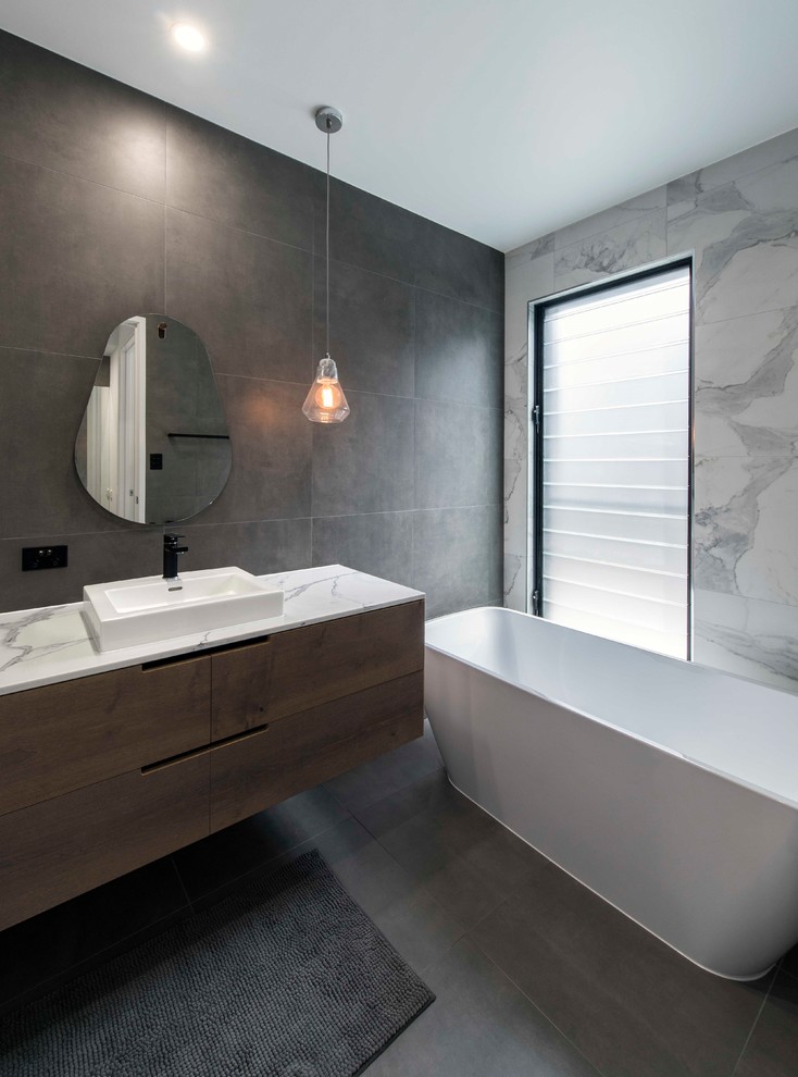 Inredning av ett modernt vit vitt badrum med dusch, med släta luckor, skåp i mellenmörkt trä, ett fristående badkar, grå kakel, vit kakel, grå väggar, ett fristående handfat och grått golv