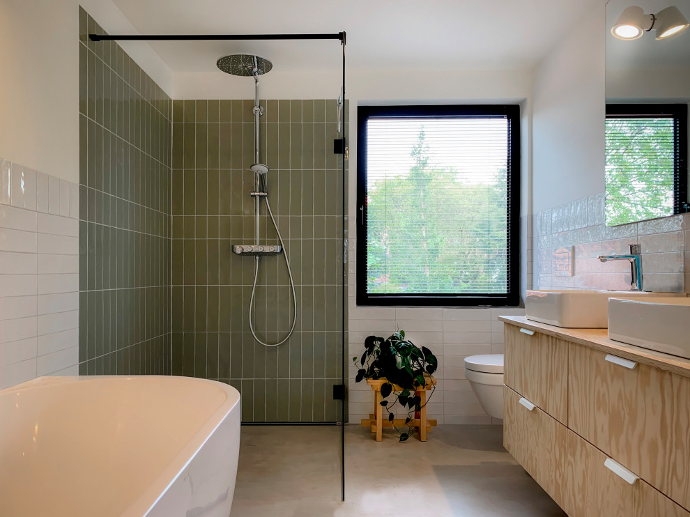 Foto på ett mellanstort funkis beige badrum för barn, med släta luckor, skåp i ljust trä, ett fristående badkar, en hörndusch, en vägghängd toalettstol, grön kakel, vita väggar, ett fristående handfat, träbänkskiva, grått golv, keramikplattor, betonggolv och med dusch som är öppen