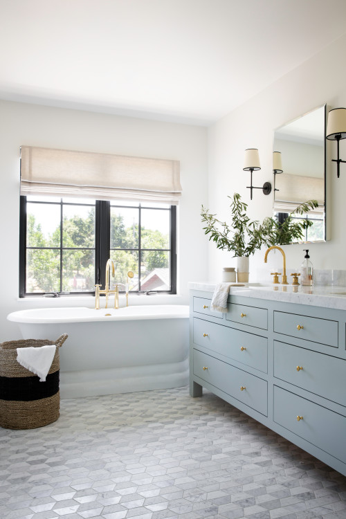 Light-blue shaker bathroom cabinets with brass hardware and marble countertop beside a freestanding tub, black-frame window, and hex tile floor.