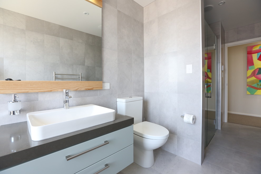 Example of a mid-sized trendy gray tile and ceramic tile ceramic tile bathroom design in Auckland with flat-panel cabinets, blue cabinets, a two-piece toilet, gray walls, a vessel sink and quartz countertops