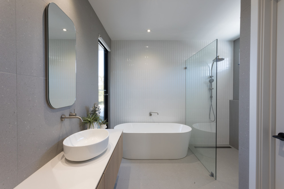 Bild på ett funkis vit vitt badrum, med skåp i ljust trä, ett fristående badkar, en öppen dusch, grå kakel, porslinskakel och kaklad bänkskiva