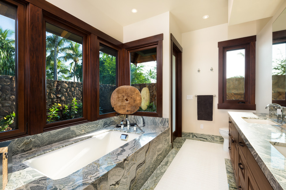 Medium sized world-inspired ensuite bathroom in Hawaii with a submerged sink, recessed-panel cabinets, dark wood cabinets, granite worktops, a submerged bath, a walk-in shower, a one-piece toilet, grey tiles, stone slabs, beige walls and marble flooring.