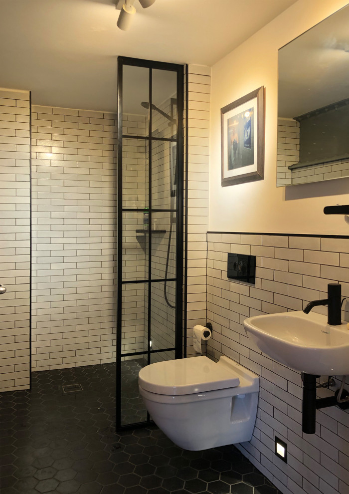 Photo of a small industrial ensuite wet room bathroom in London with a wall mounted toilet, white tiles, ceramic tiles, white walls, cement flooring, a wall-mounted sink, black floors and an open shower.