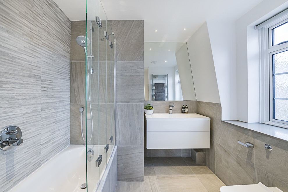 Photo of a medium sized contemporary ensuite bathroom in London with flat-panel cabinets, white cabinets, a built-in bath, a shower/bath combination, a wall mounted toilet, white walls, porcelain flooring, an integrated sink, grey floors and a hinged door.