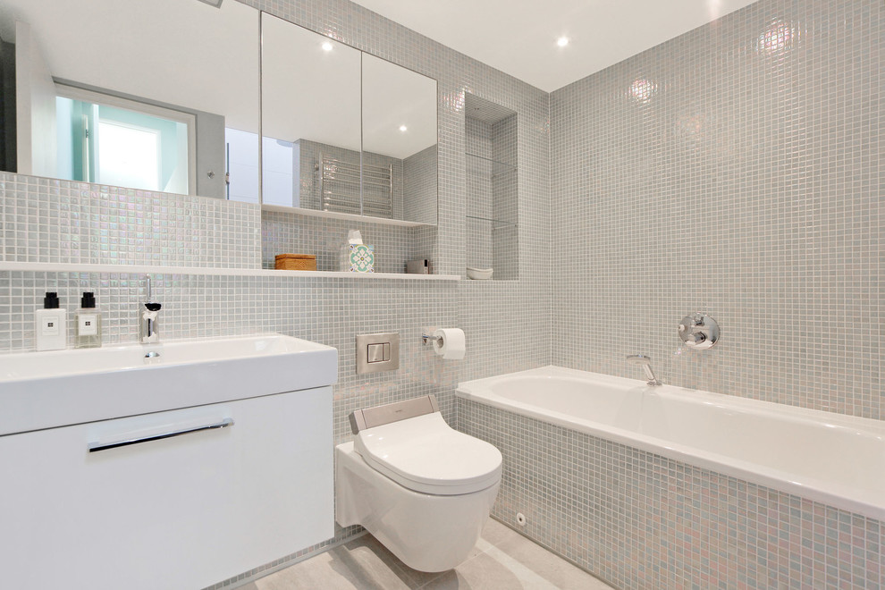 Cette image montre une salle de bain principale design de taille moyenne avec un placard à porte plane, des portes de placard blanches, une baignoire d'angle, WC suspendus, un carrelage gris, un mur gris, un sol gris et un lavabo intégré.