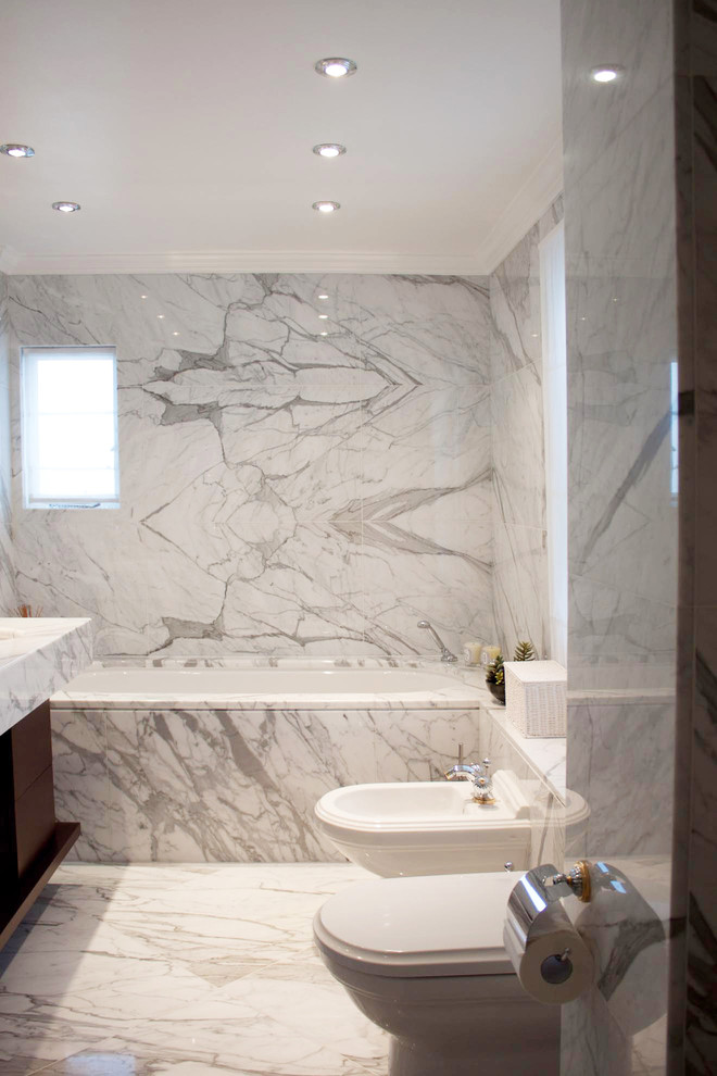 Bild på ett funkis badrum, med släta luckor, skåp i mellenmörkt trä, marmorbänkskiva, ett undermonterat badkar, en toalettstol med hel cisternkåpa, vit kakel, vita väggar, marmorgolv och marmorkakel