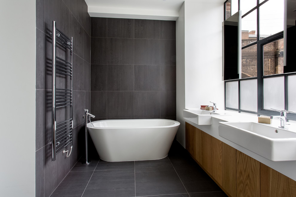 Foto på ett industriellt badrum, med ett fristående handfat, släta luckor, skåp i mellenmörkt trä, ett fristående badkar, grå kakel och vita väggar