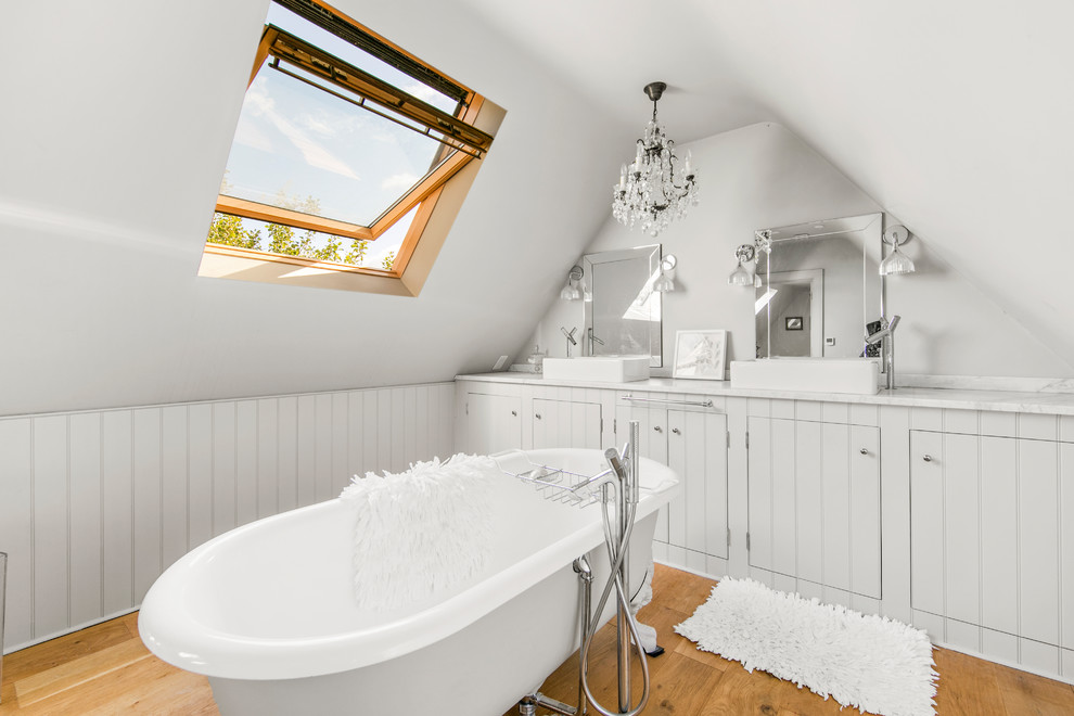 Cette image montre une salle de bain principale traditionnelle avec des portes de placard grises, une baignoire sur pieds, un mur gris, parquet clair, une vasque et un plan de toilette blanc.