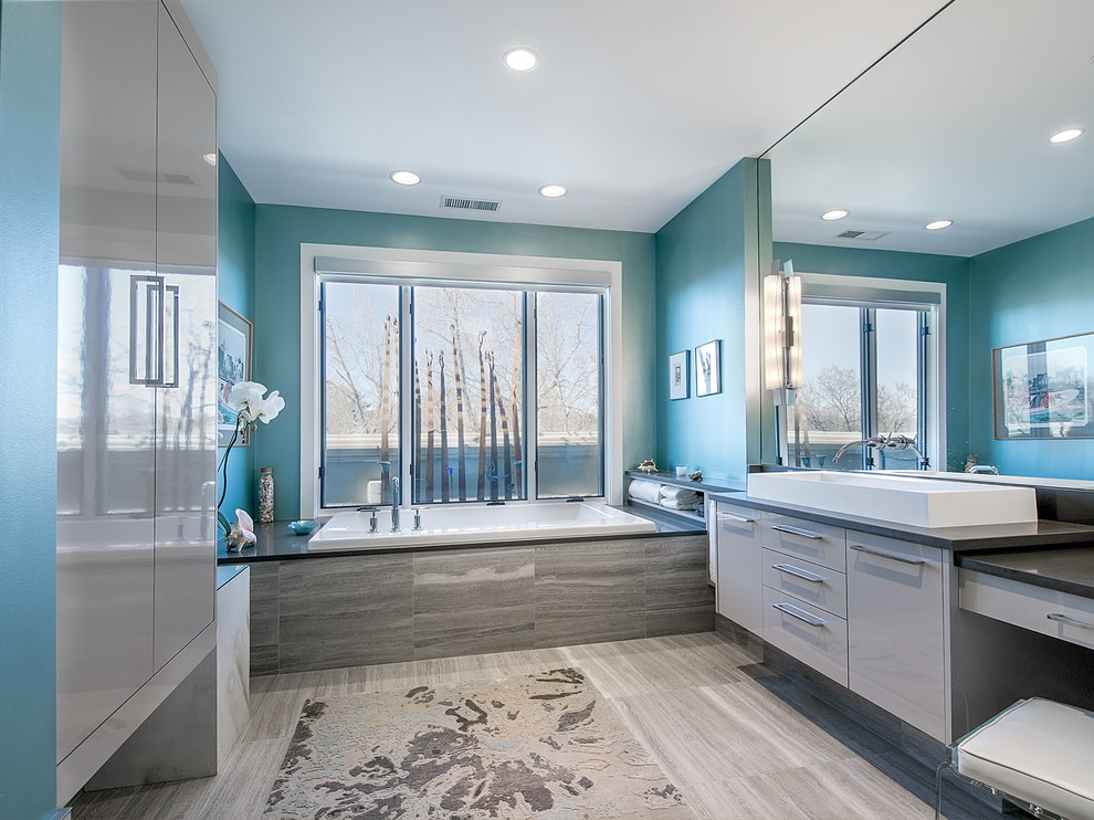 Modelo de cuarto de baño actual con lavabo sobreencimera, armarios con paneles lisos, puertas de armario grises, bañera encastrada y baldosas y/o azulejos grises