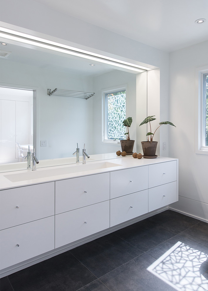 Example of a large minimalist master porcelain tile and black floor bathroom design in DC Metro with flat-panel cabinets, white cabinets, a one-piece toilet, white walls, a trough sink and solid surface countertops