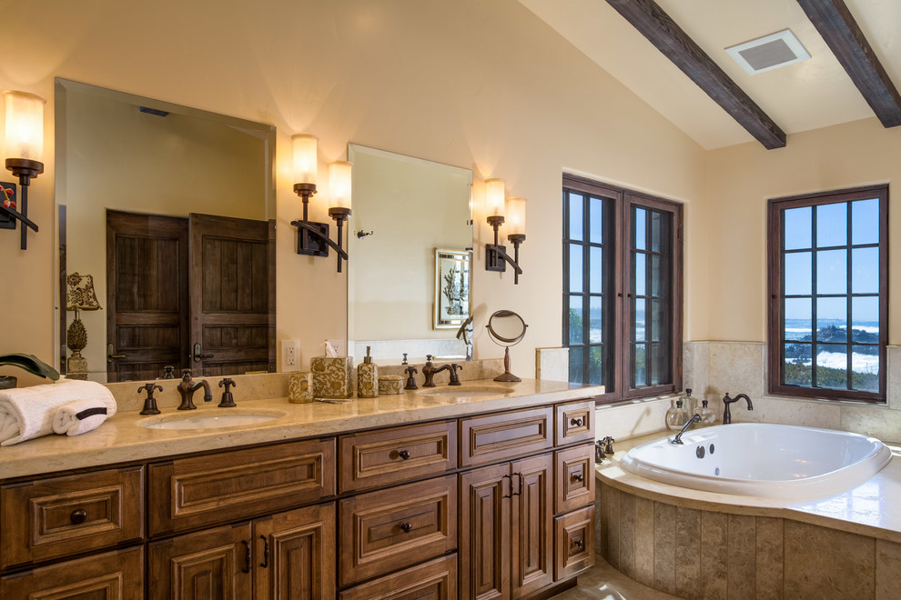 Cette image montre une grande salle de bain méditerranéenne en bois brun avec un lavabo encastré, un mur beige, un carrelage beige, une baignoire posée, une fenêtre et un placard avec porte à panneau surélevé.