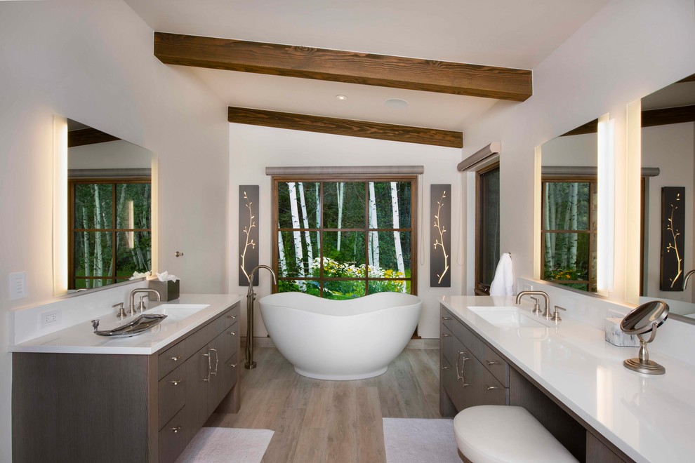 Photo of a rustic bathroom in Denver with flat-panel cabinets, grey cabinets, a freestanding bath, white walls, medium hardwood flooring, a submerged sink, brown floors and white worktops.