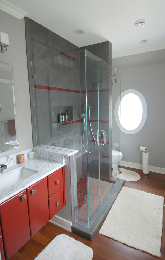Пример оригинального дизайна: ванная комната среднего размера в стиле неоклассика (современная классика) с плоскими фасадами, красными фасадами, открытым душем, унитазом-моноблоком, серой плиткой, керамогранитной плиткой, серыми стенами, паркетным полом среднего тона, душевой кабиной, врезной раковиной и мраморной столешницей