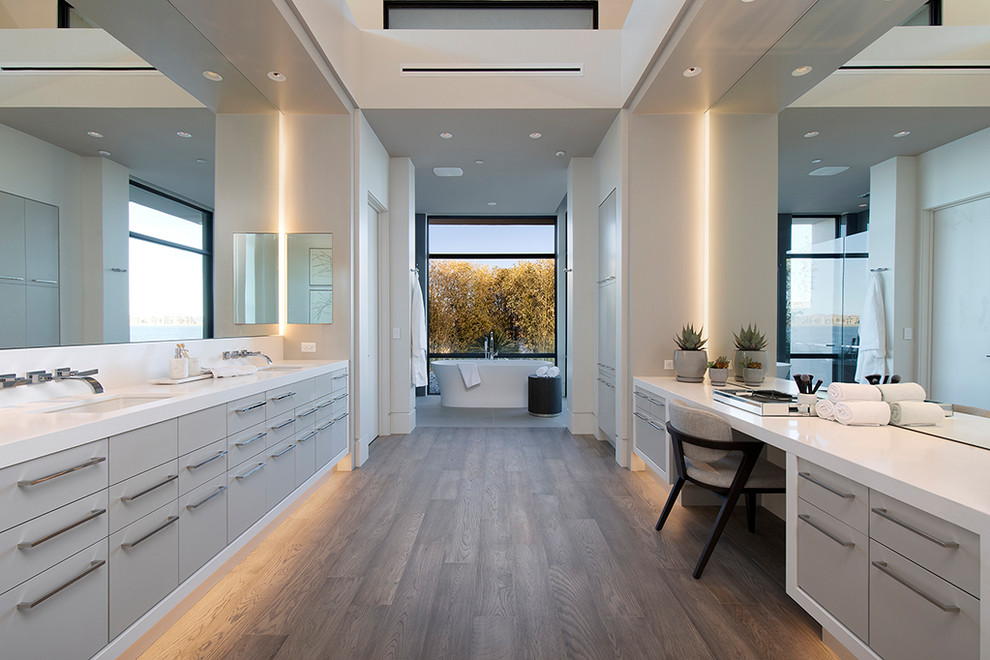 Пример оригинального дизайна: главная ванная комната в современном стиле с плоскими фасадами, серыми фасадами, отдельно стоящей ванной, белыми стенами, темным паркетным полом, врезной раковиной, коричневым полом, белой столешницей и зеркалом с подсветкой