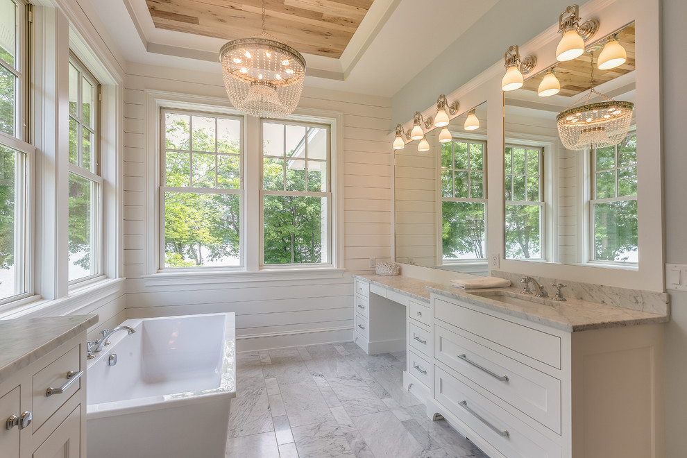 Cette photo montre une salle de bain bord de mer avec un placard à porte shaker, des portes de placard beiges, une baignoire indépendante, un mur blanc, un sol en marbre, un lavabo encastré, un plan de toilette en marbre et un sol gris.