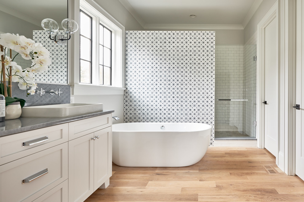 Lantlig inredning av ett en-suite badrum, med skåp i shakerstil, vita skåp, ett fristående badkar, en dusch i en alkov, flerfärgad kakel, grå väggar, ljust trägolv, ett fristående handfat, beiget golv och dusch med gångjärnsdörr