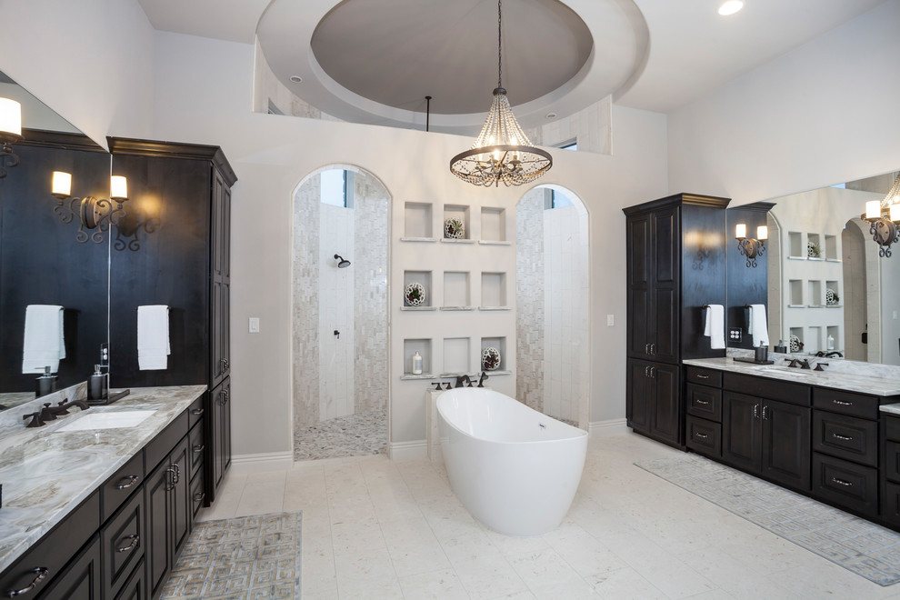 This is an example of a large traditional ensuite bathroom in Austin with a freestanding bath, a built-in shower, a two-piece toilet, marble tiles, limestone flooring, a submerged sink, grey worktops, raised-panel cabinets, dark wood cabinets, beige tiles, white walls, quartz worktops, beige floors and an open shower.