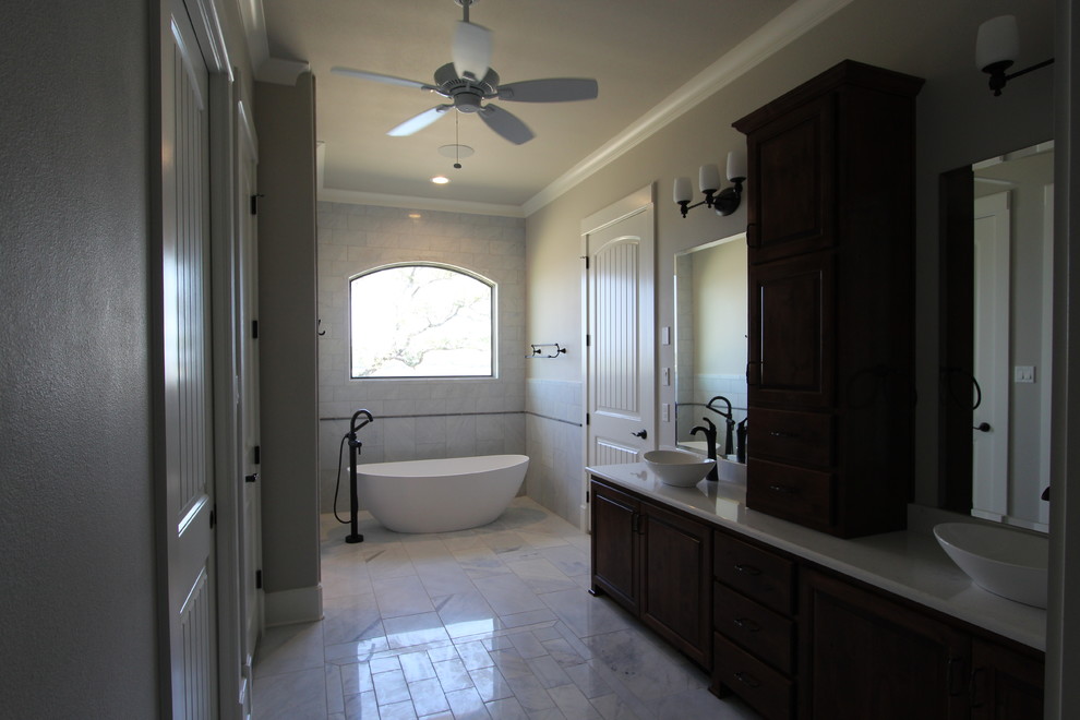 Large traditional ensuite bathroom in Austin with raised-panel cabinets, dark wood cabinets, a freestanding bath, a corner shower, a two-piece toilet, white tiles, marble tiles, a vessel sink, marble worktops, a hinged door, beige walls, marble flooring and white floors.