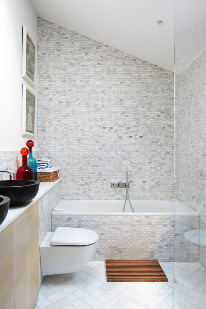 Kleines Modernes Badezimmer mit flächenbündigen Schrankfronten, hellen Holzschränken, Einbaubadewanne, offener Dusche, Toilette mit Aufsatzspülkasten, grauen Fliesen, Keramikfliesen, weißer Wandfarbe, Mosaik-Bodenfliesen, Waschtischkonsole und Quarzwerkstein-Waschtisch in London