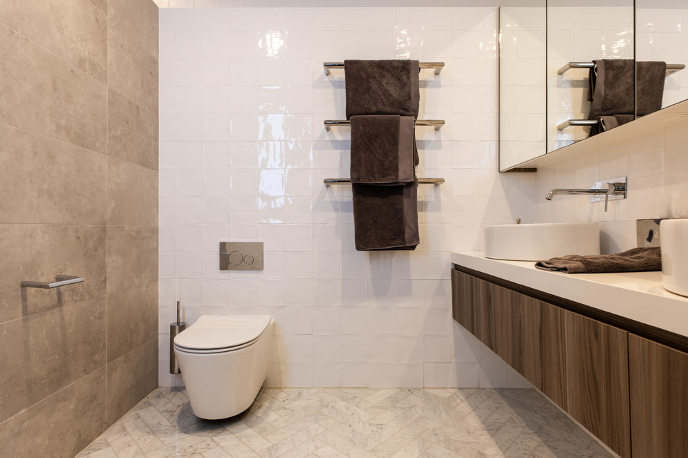 Cette photo montre une salle de bain tendance en bois brun avec un placard à porte plane, WC suspendus, un carrelage beige, un carrelage marron, un carrelage blanc, un mur blanc, une vasque, un sol gris et un plan de toilette blanc.