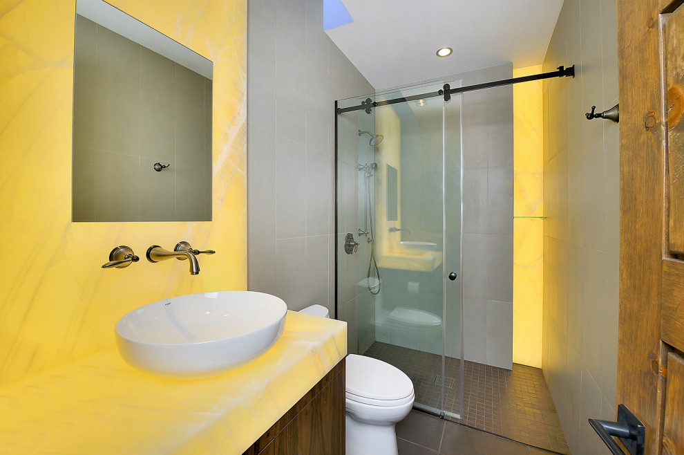 На фото: ванная комната среднего размера в стиле модернизм с плоскими фасадами, темными деревянными фасадами, душем в нише, унитазом-моноблоком, серой плиткой, цементной плиткой, душевой кабиной, настольной раковиной, столешницей из оникса, коричневым полом, душем с раздвижными дверями и желтой столешницей