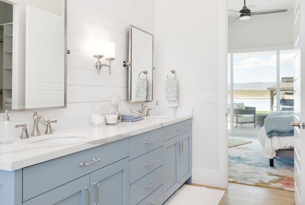 Cette photo montre une salle de bain bord de mer avec un placard à porte shaker, des portes de placard bleues, un mur blanc, un sol en bois brun, un lavabo encastré, un sol marron, un plan de toilette blanc, meuble double vasque et meuble-lavabo encastré.