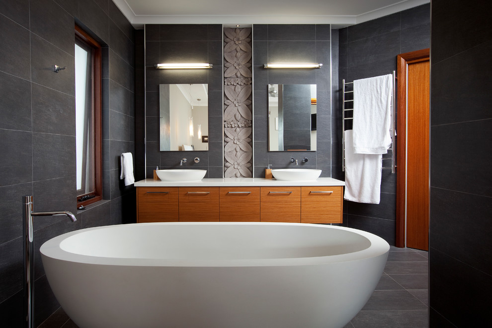 На фото: ванная комната среднего размера в современном стиле с настольной раковиной, фасадами цвета дерева среднего тона, столешницей из искусственного кварца, отдельно стоящей ванной, серой плиткой и плоскими фасадами