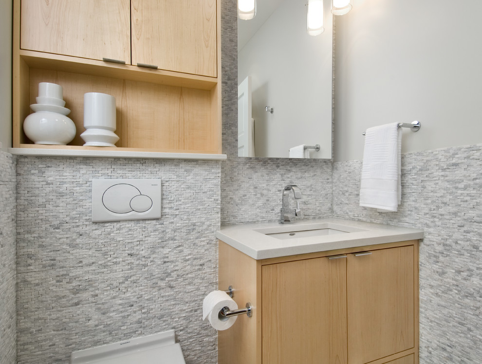 Bild på ett litet funkis badrum, med ett undermonterad handfat, släta luckor, skåp i ljust trä, bänkskiva i kvarts, en vägghängd toalettstol, vit kakel, stenkakel, grå väggar och klinkergolv i porslin