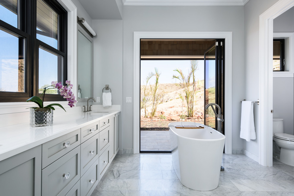 This is an example of a large traditional ensuite bathroom in Hawaii with shaker cabinets, grey cabinets, a freestanding bath, a one-piece toilet, grey walls, marble flooring, a submerged sink and engineered stone worktops.