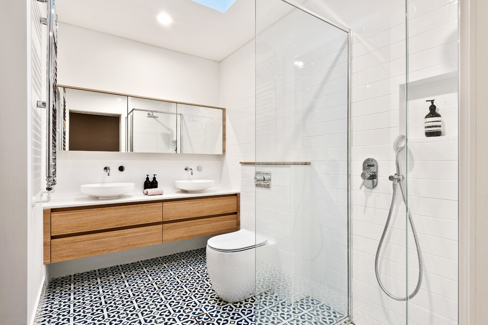 Lantlig inredning av ett vit vitt badrum, med släta luckor, skåp i mellenmörkt trä, en kantlös dusch, en toalettstol med hel cisternkåpa, vit kakel, vita väggar, cementgolv, ett fristående handfat, flerfärgat golv och med dusch som är öppen