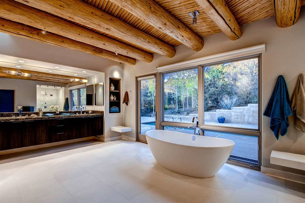 Cette image montre une très grande salle de bain principale sud-ouest américain en bois foncé avec un placard à porte plane, une baignoire indépendante, un mur blanc, un sol en carrelage de porcelaine, un lavabo encastré et une fenêtre.