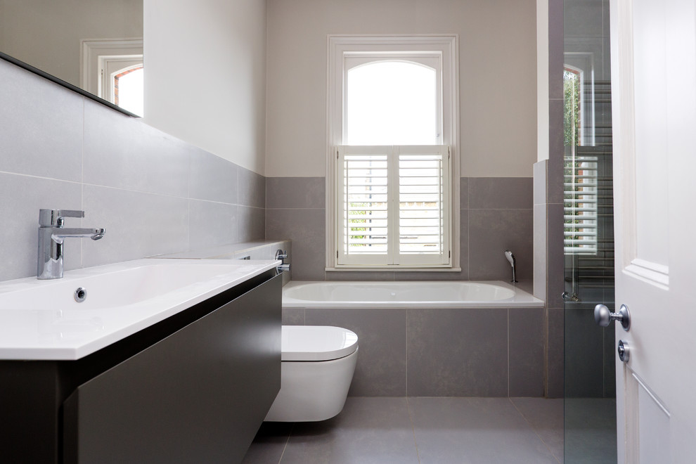 Idée de décoration pour une salle d'eau design avec un placard à porte plane, des portes de placard noires, une baignoire posée, WC suspendus, un carrelage gris, un mur blanc, un lavabo intégré, un sol gris et un plan de toilette blanc.