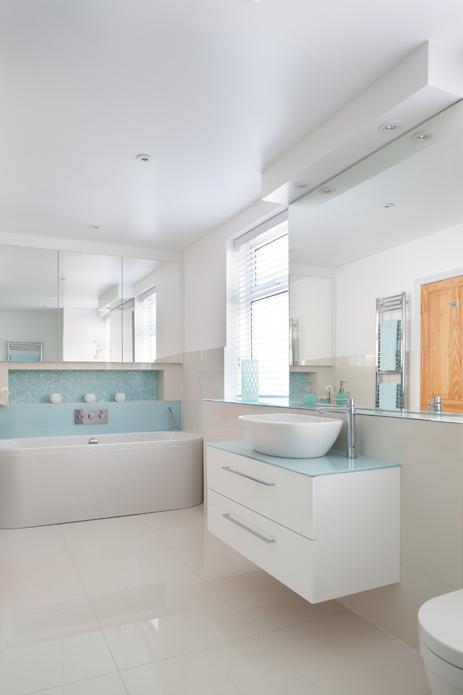 他の地域にある低価格の中くらいなコンテンポラリースタイルのおしゃれなお風呂の窓 (ベッセル式洗面器、ガラスの洗面台、置き型浴槽、壁掛け式トイレ、白いタイル、青いタイル、磁器タイルの床、フラットパネル扉のキャビネット、白いキャビネット、白い壁、青い洗面カウンター) の写真