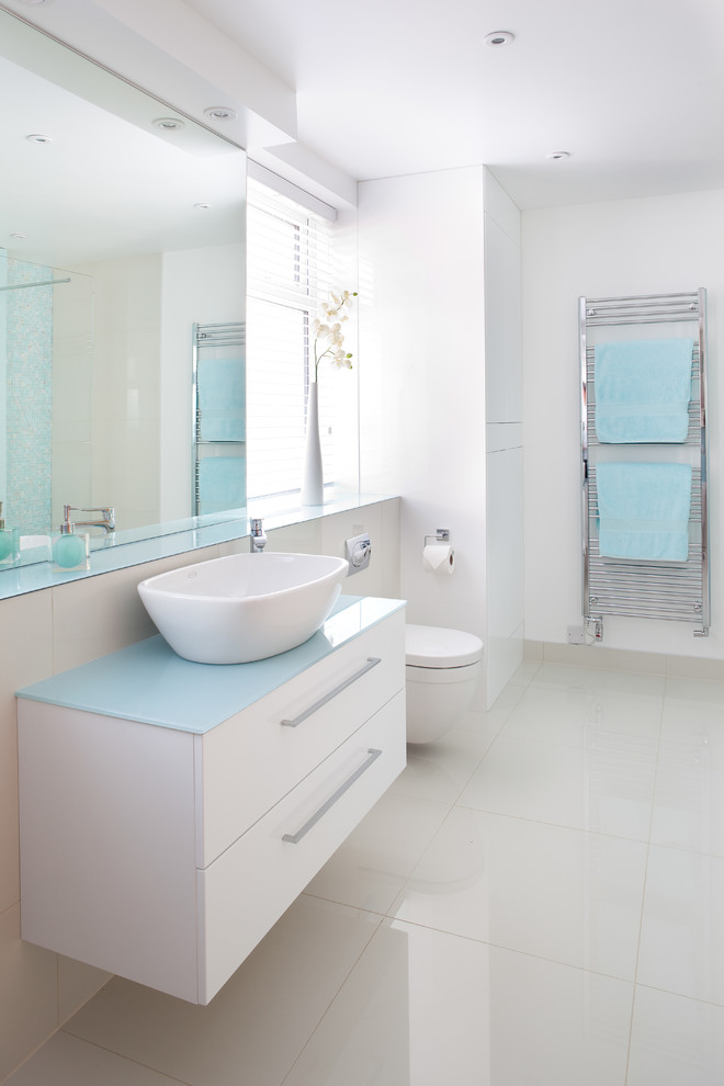 Imagen de cuarto de baño contemporáneo de tamaño medio con lavabo tipo consola, encimera de vidrio, sanitario de pared, baldosas y/o azulejos blancos y suelo de baldosas de porcelana