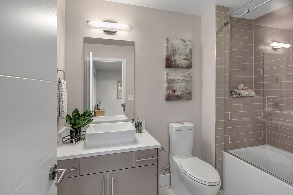 Foto på ett funkis vit badrum, med släta luckor, grå skåp, ett badkar i en alkov, en toalettstol med separat cisternkåpa, grå kakel, tunnelbanekakel, grå väggar och ett fristående handfat