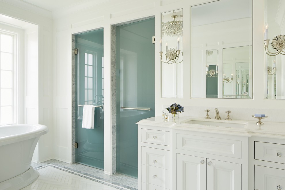 Klassisk inredning av ett stort en-suite badrum, med ett undermonterad handfat, luckor med infälld panel, vita skåp, marmorbänkskiva, ett fristående badkar, en kantlös dusch, en toalettstol med separat cisternkåpa, vit kakel, stenkakel, vita väggar och marmorgolv