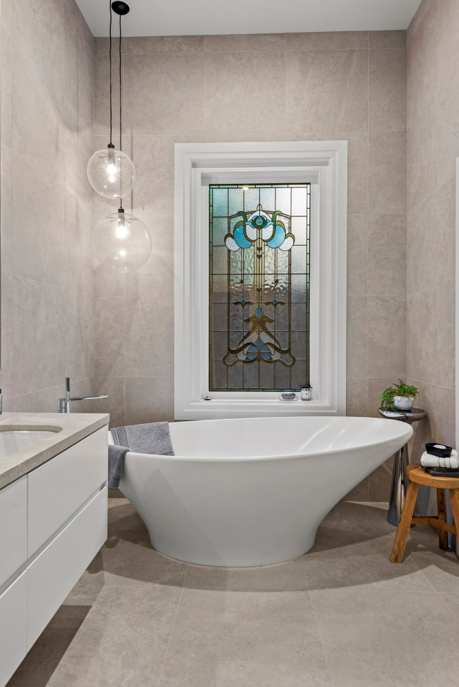 Bild på ett mellanstort vintage beige beige badrum med dusch, med luckor med lamellpanel, vita skåp, ett fristående badkar, grå kakel, keramikplattor, grå väggar, cementgolv, ett undermonterad handfat, bänkskiva i kalksten och beiget golv
