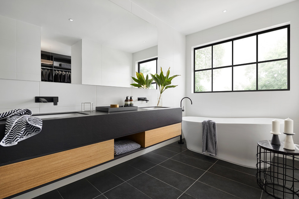 Cette image montre une salle d'eau design avec un placard à porte plane, des portes de placard noires, une baignoire indépendante, un carrelage blanc, un mur blanc, un lavabo encastré et un sol noir.
