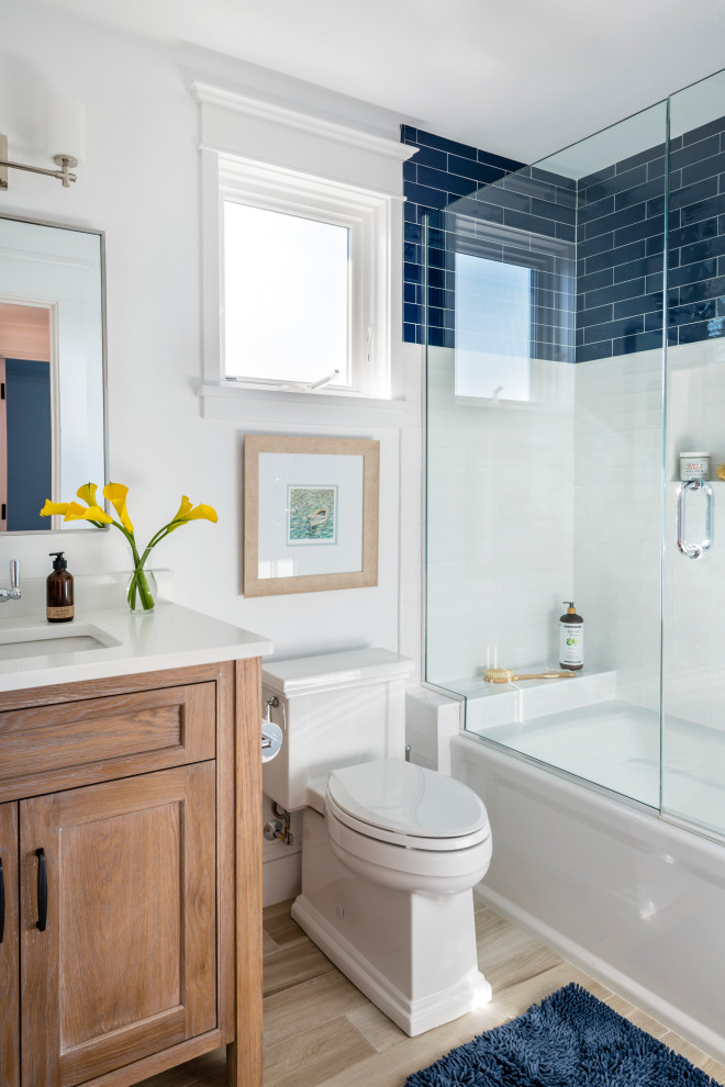 Exemple d'une salle de bain bord de mer en bois brun avec un placard à porte shaker, une baignoire en alcôve, un combiné douche/baignoire, un carrelage bleu, un carrelage blanc, un mur blanc, un lavabo encastré, un sol beige, une cabine de douche à porte battante, un plan de toilette blanc, meuble-lavabo sur pied et meuble simple vasque.